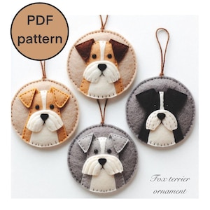 May include: Four felt ornaments shaped like dog heads. The ornaments are brown, tan, grey, and white. Each ornament has a loop for hanging. The text "Fox terrier ornament" is at the bottom of the image.