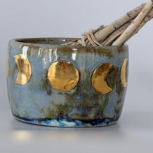 May include: A ceramic bowl with a blue and green mottled glaze featuring gold-coloured moon phase decals around the exterior. A bundle of dried, light-coloured wooden sticks is placed inside the bowl, extending over the top rim of the vessel.