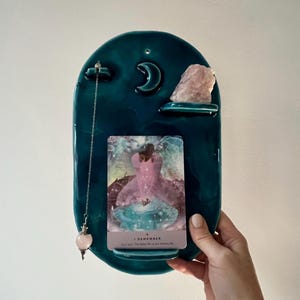 Pode incluir: Suporte de parede em cerâmica azul-turquesa com uma lua crescente, uma prateleira de nuvem e um cristal de quartzo rosa. Um pêndulo pende do topo e uma carta de tarô é exibida no centro. A carta diz "I Remember".