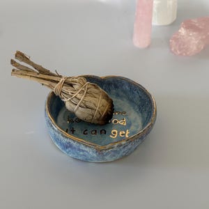 May include: A heart-shaped blue ceramic dish with a gold rim and gold lettering that reads "how good it can get" on the interior base. A dried sage bundle is resting inside the dish, accompanied by three crystal points in the background.