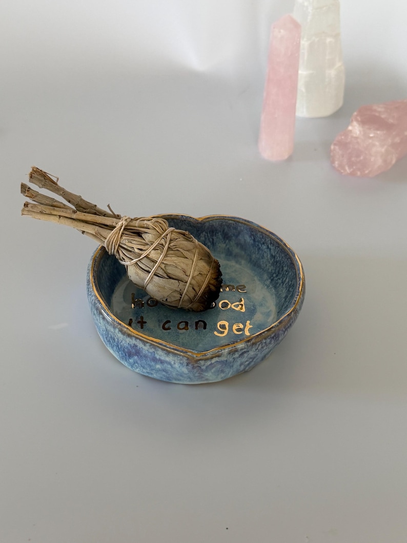 May include: A heart-shaped blue ceramic dish with a gold rim and gold lettering that reads "how good it can get" on the interior base. A dried sage bundle is resting inside the dish, accompanied by three crystal points in the background.