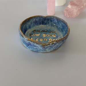 May include: A heart-shaped ceramic trinket dish with a mottled blue and white glaze and a gold-painted rim. The interior features the gold-lettered text, "Show me how good it can get," centered on the base of the bowl.