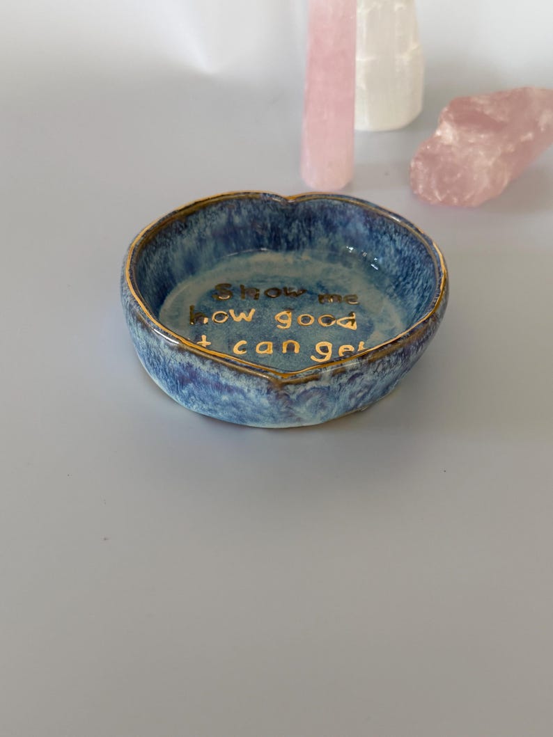 May include: A heart-shaped ceramic trinket dish with a mottled blue and white glaze and a gold-painted rim. The interior features the gold-lettered text, "Show me how good it can get," centered on the base of the bowl.