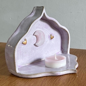 May include: A lavender ceramic candle holder with a curved, arch-like back featuring a central pale pink crescent moon accent and two small gold-toned heart embellishments. A single white tea light candle sits on the flat base of the holder.