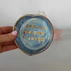 May include: A heart-shaped ceramic trinket dish with a mottled blue glaze and a gold-painted rim. The center of the dish features the gold-lettered text, Show me how good it can get, written in a simple, lowercase, sans-serif font style.