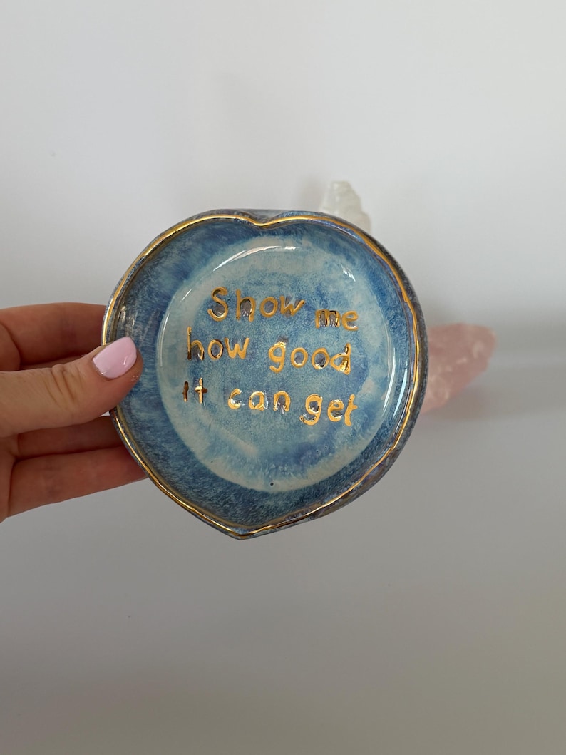 May include: A heart-shaped ceramic trinket dish with a mottled blue glaze and a gold-painted rim. The center of the dish features the gold-lettered text, Show me how good it can get, written in a simple, lowercase, sans-serif font style.