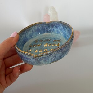 May include: A small, round, blue ceramic trinket dish with a mottled, textured glaze and a gold-painted rim. The interior features the gold-lettered text "Show me how good it can be" inscribed in the center of the bowl's basin.