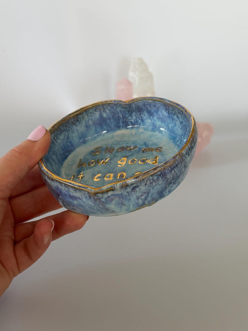 May include: A small, round, blue ceramic trinket dish with a mottled, textured glaze and a gold-painted rim. The interior features the gold-lettered text "Show me how good it can be" inscribed in the center of the bowl's basin.