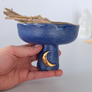 Może przedstawiać: Głęboko niebieska ceramiczna miska na nóżce o teksturowanym wykończeniu ze złotym półksiężycem namalowanym na podstawie. Krawędź miski jest podkreślona złotą farbą i zawiera pęczek wysuszonych drewnianych patyczków związanych sznurkiem.