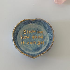 May include: A heart-shaped ceramic trinket dish with a mottled blue glaze and a gold-painted rim. The interior features the gold-lettered text "Show me how good it can get" centered in the bowl. The dish has a smooth, glossy finish throughout.
