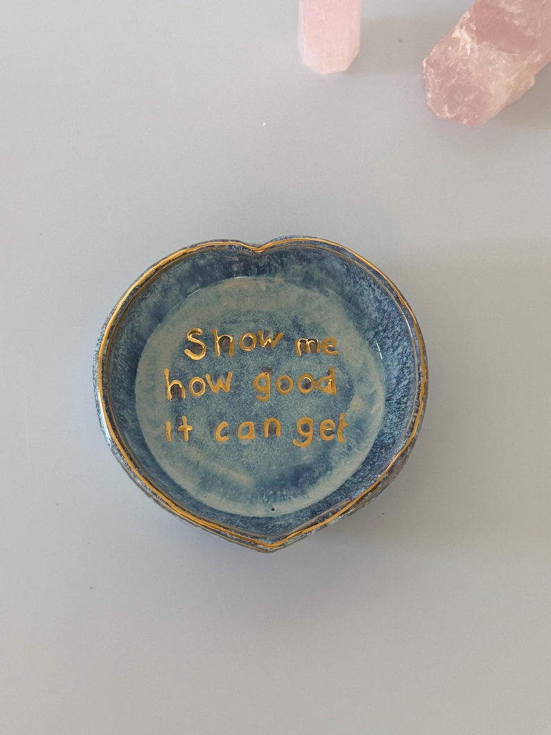 May include: A heart-shaped ceramic trinket dish with a mottled blue glaze and a gold-painted rim. The interior features the gold-lettered text "Show me how good it can get" centered in the bowl. The dish has a smooth, glossy finish throughout.