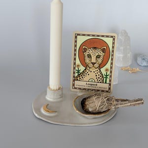 May include: A ceramic tray with a gold crescent moon accent, holding a tall off-white taper candle, a small bowl with a sage smudge stick, and a rectangular card featuring a leopard illustration with the text Leopard, Strength, Patience, Instinct.