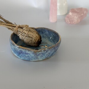 May include: A small, round, blue ceramic bowl with a gold-trimmed rim and a sage smudge stick resting inside. The interior of the bowl features gold lettering. In the background, there are three light-colored, translucent crystal formations.
