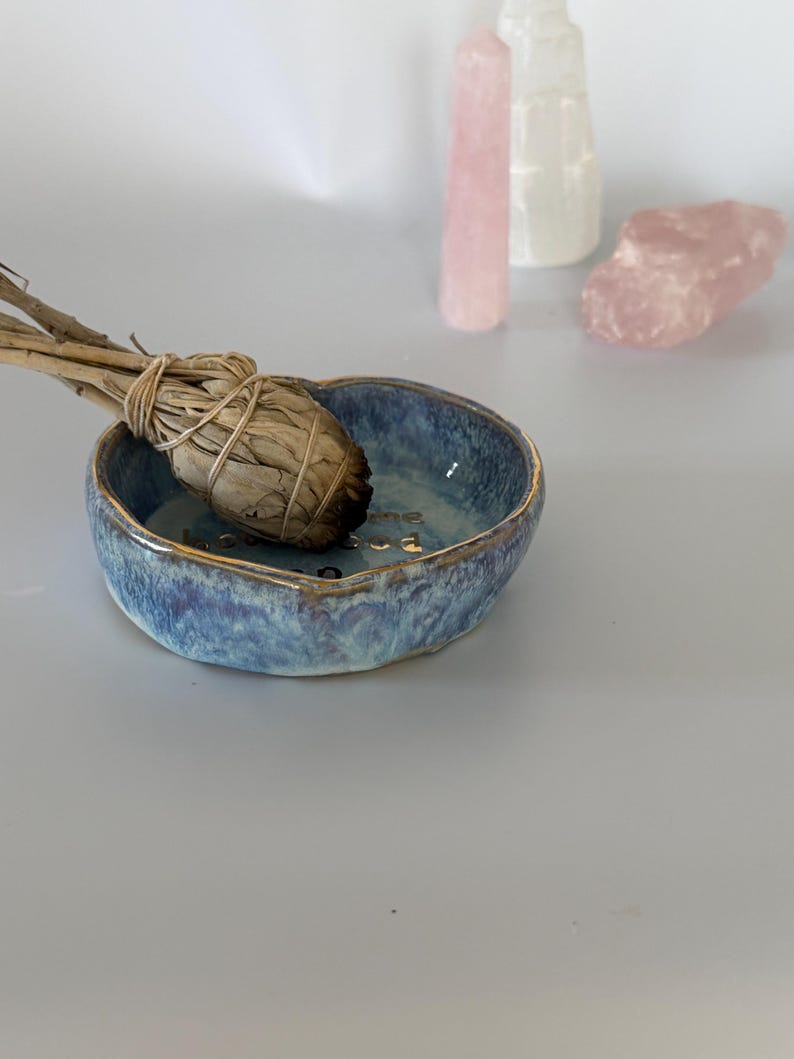 May include: A small, round, blue ceramic bowl with a gold-trimmed rim and a sage smudge stick resting inside. The interior of the bowl features gold lettering. In the background, there are three light-colored, translucent crystal formations.