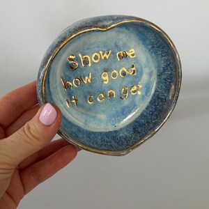 May include: A heart-shaped ceramic trinket dish with a blue and white mottled glaze. The interior features the gold-colored text "Show me how good it can get" written in a cursive font. The rim of the dish is finished with a thin gold-colored trim.