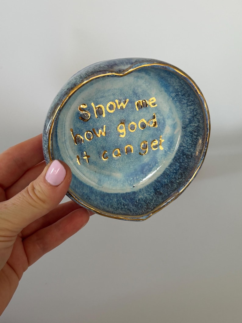 May include: A heart-shaped ceramic trinket dish with a blue and white mottled glaze. The interior features the gold-colored text "Show me how good it can get" written in a cursive font. The rim of the dish is finished with a thin gold-colored trim.
