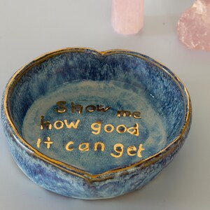 May include: A heart-shaped ceramic trinket dish with a blue mottled glaze and a gold-painted rim. The interior features the gold-lettered text "Show me how good it can get" centered on the base, with a glossy finish throughout the piece.