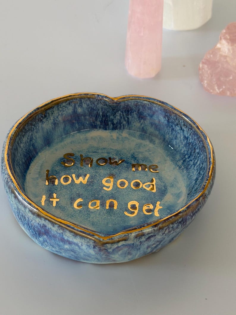 May include: A heart-shaped ceramic trinket dish with a blue mottled glaze and a gold-painted rim. The interior features the gold-lettered text "Show me how good it can get" centered on the base, with a glossy finish throughout the piece.