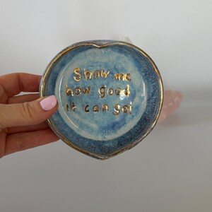 May include: A heart-shaped ceramic trinket dish with a blue mottled glaze and a gold-painted rim. The center features the gold-lettered text "Show me how good it can get" written in a cursive script, adding a metallic detail to the blue surface.