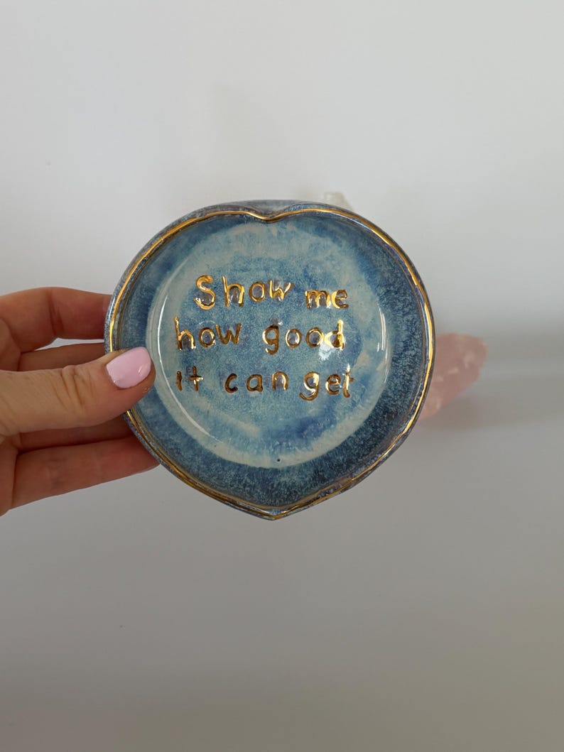 May include: A heart-shaped ceramic trinket dish with a blue mottled glaze and a gold-painted rim. The center features the gold-lettered text "Show me how good it can get" written in a cursive script, adding a metallic detail to the blue surface.