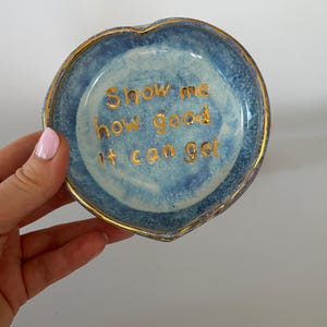 May include: A heart-shaped ceramic trinket dish with a mottled blue glaze and a gold-painted rim. The center features the gold-lettered text "Show me how good it can get" written in a simple, sans-serif font against the blue background.