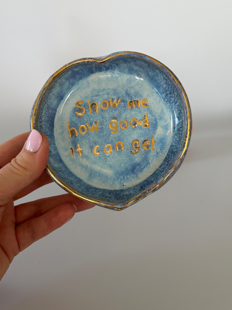 May include: A heart-shaped ceramic trinket dish with a mottled blue glaze and a gold-painted rim. The center features the gold-lettered text "Show me how good it can get" written in a simple, sans-serif font against the blue background.