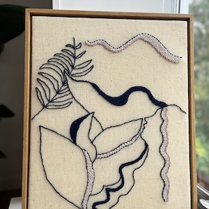 May include: Embroidered artwork in a wooden frame. The piece features a cream background with dark blue and light beige embroidered floral and abstract designs. A fern and a flower are visible.