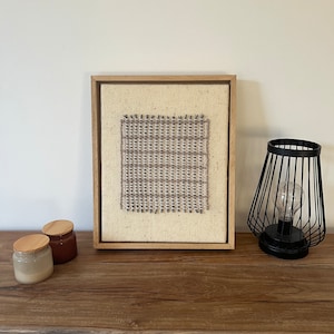 May include: A framed woven textile art piece with a natural wood frame. The artwork features a rectangular woven design in shades of gray and beige. A black metal wire lamp and two small jars with wooden lids are on a wooden table.