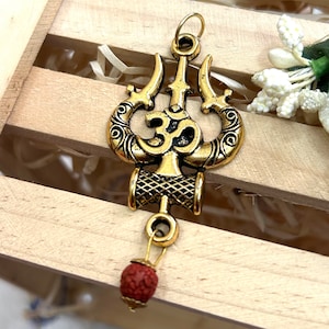 May include: Gold-toned metal pendant featuring a trident with an Om symbol in the centre, with a red bead hanging from the bottom.