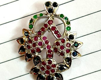 May include: A silver pendant with a heart-shaped design, featuring a cluster of red, green, and blue gemstones. The pendant is intricate and detailed, with a delicate floral motif.