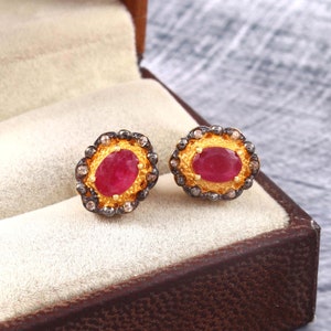 Op de afbeelding: Een paar gouden oorbellen met een rode ovale edelsteen omringd door een halo van kleinere diamanten. De oorbellen hebben een zwarte geoxideerde afwerking.