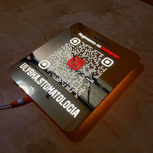 Puede incluir: Un letrero rectangular iluminado con un código QR y el logotipo de Instagram. El letrero tiene el texto "Ulybka.Stomatologia" y "Подпишись на Instagram" en blanco. Está enchufado con un cable rojo y azul.