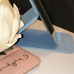 May include: A light blue foldable mirror with a pink case that says "Lyra" on it. The mirror is propped up at an angle.