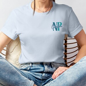 May include: A light blue t-shirt with a teal and blue monogram of the letters "NP" and the name "Christina" written in cursive.