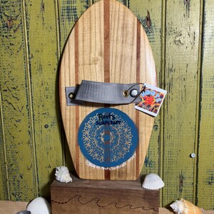 May include: Wooden surfboard-shaped wall decor with a grey strap and a blue and gold circular logo. The surfboard is displayed on a wooden stand with seashells. The text on the logo reads "Roots Surfcraft".
