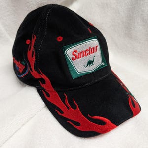 May include: Black baseball cap with red flame embroidery. Features a Sinclair logo patch with a green border, white background, and a dinosaur silhouette. The cap has a curved brim and a structured crown.