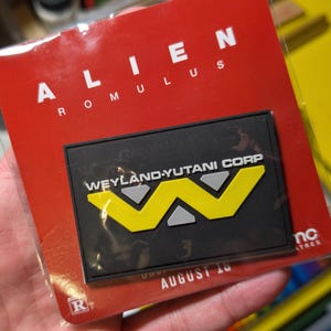 May include: A rectangular black patch featuring the yellow and grey Weyland-Yutani Corp logo, mounted on a red backing card with the white text Alien Romulus and the AMC Theatres logo. The entire item is enclosed in a clear protective plastic sleeve.