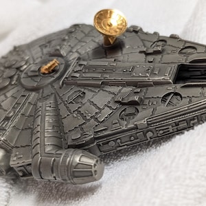 May include: A detailed silver-colored metal model of the Millennium Falcon spaceship from Star Wars. The model has a gold-colored antenna on top.