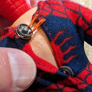 Peut inclure: Gros plan sur une figurine d'action Spiderman rouge et bleue avec une combinaison rouge et bleue. La figurine a un bouton noir sur le devant de la combinaison et le texte "MADE IN HONG KONG" est visible sur le torse de la figurine.