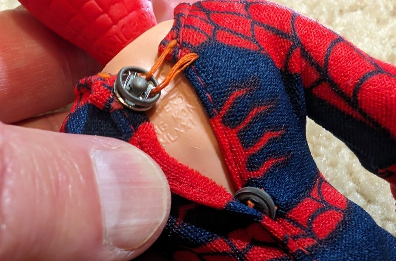 Peut inclure: Gros plan sur une figurine d'action Spiderman rouge et bleue avec une combinaison rouge et bleue. La figurine a un bouton noir sur le devant de la combinaison et le texte "MADE IN HONG KONG" est visible sur le torse de la figurine.