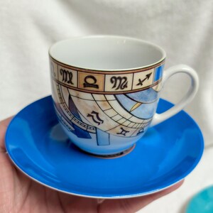 May include: A white teacup with a handle and a matching blue saucer. The teacup features a detailed design of zodiac symbols and a celestial map in shades of brown, blue, and beige. The saucer is a solid, bright blue.