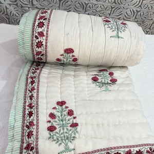 May include: A white cotton quilt with a floral print in shades of pink and green. The quilt has a quilted pattern and a border with a repeating floral design.