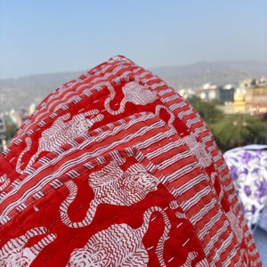 Może przedstawiać: Czerwony pikowany koc z białym nadrukiem tygrysa. Koc ma pasiasty wzór na krawędziach.