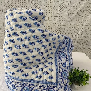 May include: A white cotton quilt with a blue floral pattern and a blue paisley border. The quilt is folded and displayed on a white surface.