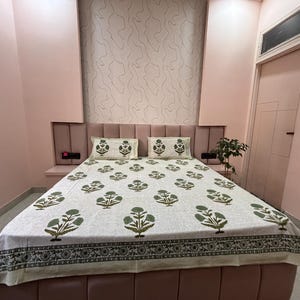 May include: A bed with a floral patterned bedspread and two matching pillowcases. The bedspread features a repeating pattern of green floral designs on a white background. The room has pink walls and a matching headboard.