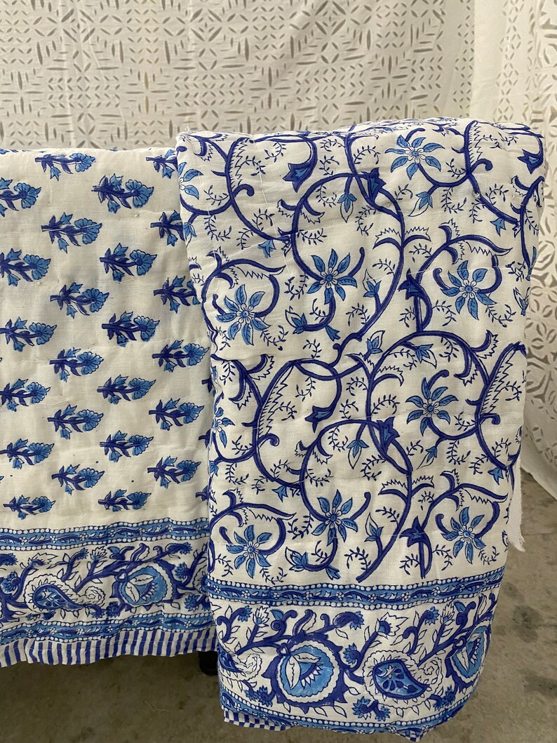 May include: Two white cotton fabric panels with blue floral patterns. The panel on the left has a smaller, repeating pattern of blue leaves and flowers. The panel on the right has a larger, more intricate pattern of blue flowers and vines. Both panels have a blue paisley border.