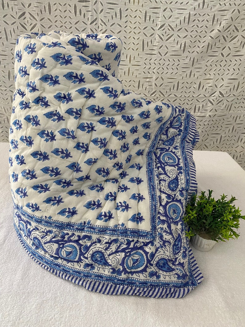 May include: A white cotton quilt with a blue floral pattern and a blue paisley border. The quilt is folded and displayed on a white surface.