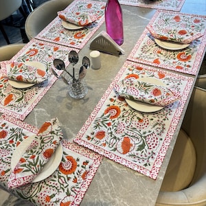 Può includere: Una tavola apparecchiata con piatti bianchi, tovaglioli floreali e tovagliette con disegni arancioni e verdi. Sono presenti anche una bottiglia di vetro rosa e posate. La tavola è apparecchiata per un pasto.