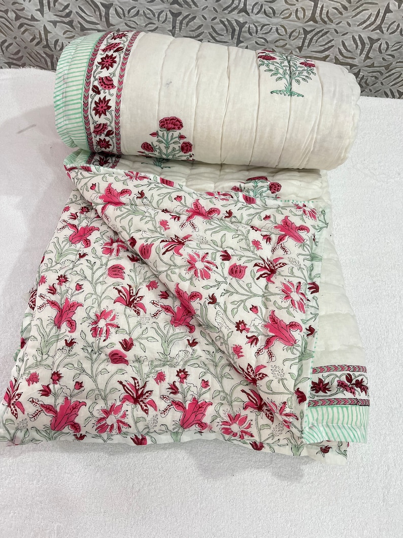 May include: A white cotton quilt with a floral print in shades of pink and green. The quilt is folded in half and has a border with a green and white striped pattern.