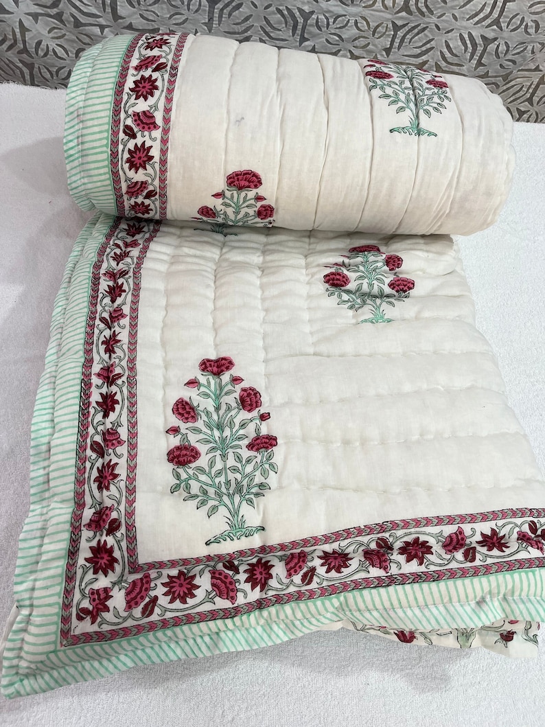 May include: A white cotton quilt with a floral print in shades of pink and green. The quilt has a quilted pattern and a border with a repeating floral design.